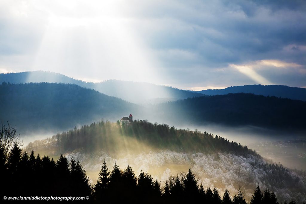 Sunrays bursting through the clouds and illuminating the church of Saint Joseph and surrounding frost, seen from the hilltop church of Saint Ana on the Ljubljana Moors in Slovenia.